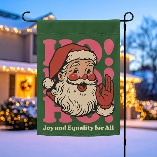 Green garden flag with a vintage Santa illustration and the phrase “Joy and Equality for All,” hanging on a metal stand in front of a warmly lit home.