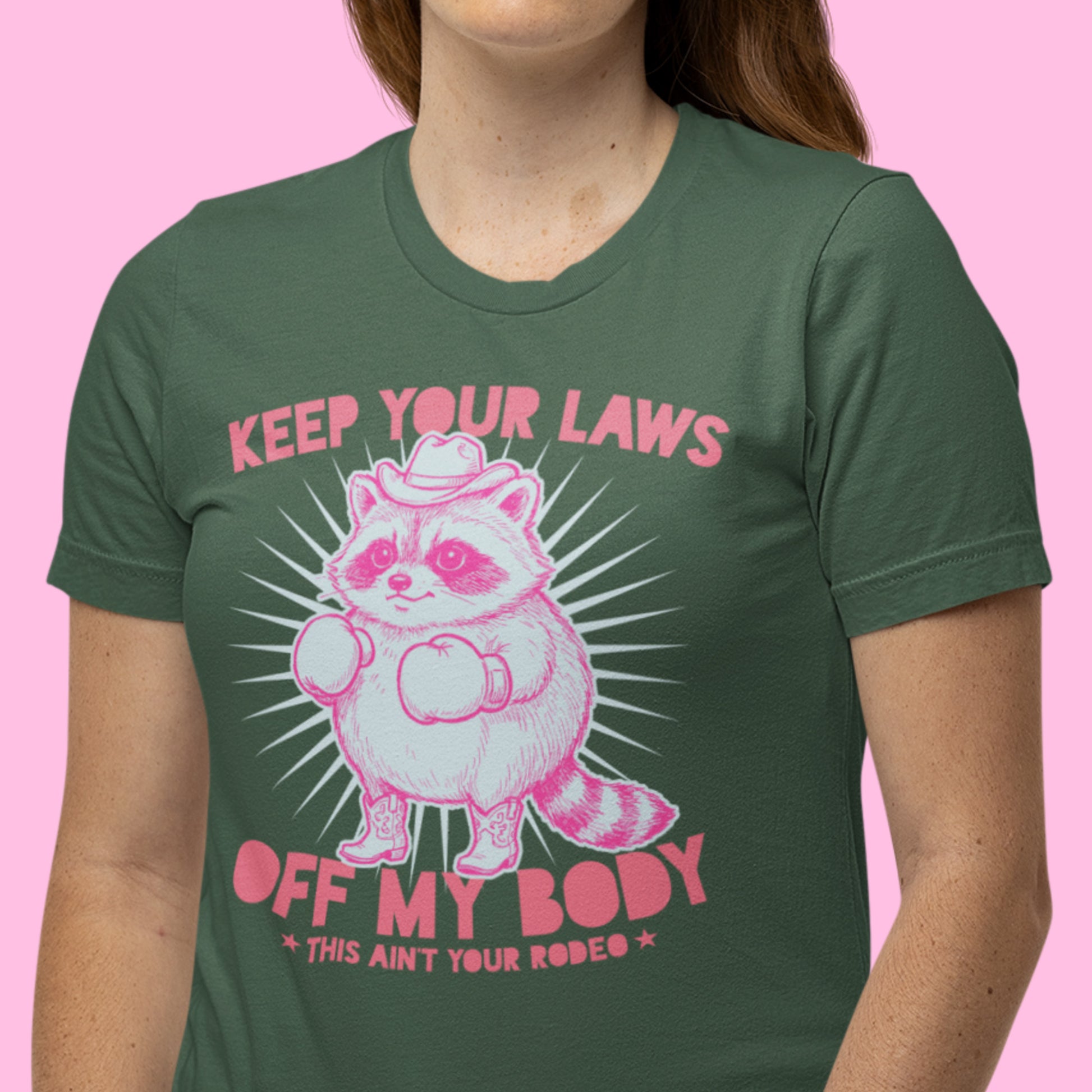 Close-up of green t-shirt featuring pink cowboy raccoon with boxing gloves and “Keep Your Laws Off My Body” western feminist protest graphic design.