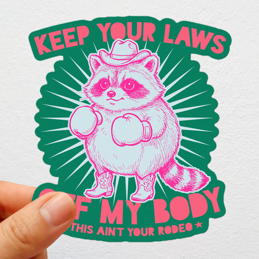 Die-cut sticker featuring pink cowboy raccoon with boxing gloves and “Keep Your Laws Off My Body” text plus “This Ain’t Your Rodeo,” bold feminist activist design.