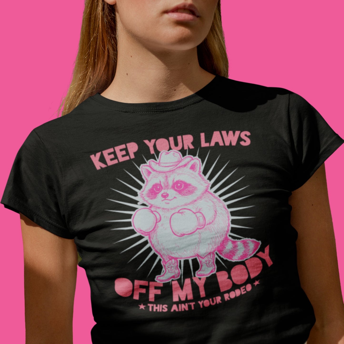 woman wearing black feminist graphic t-shirt featuring pink cowboy raccoon illustration with “Keep Your Laws Off My Body” slogan against bright pink background.
