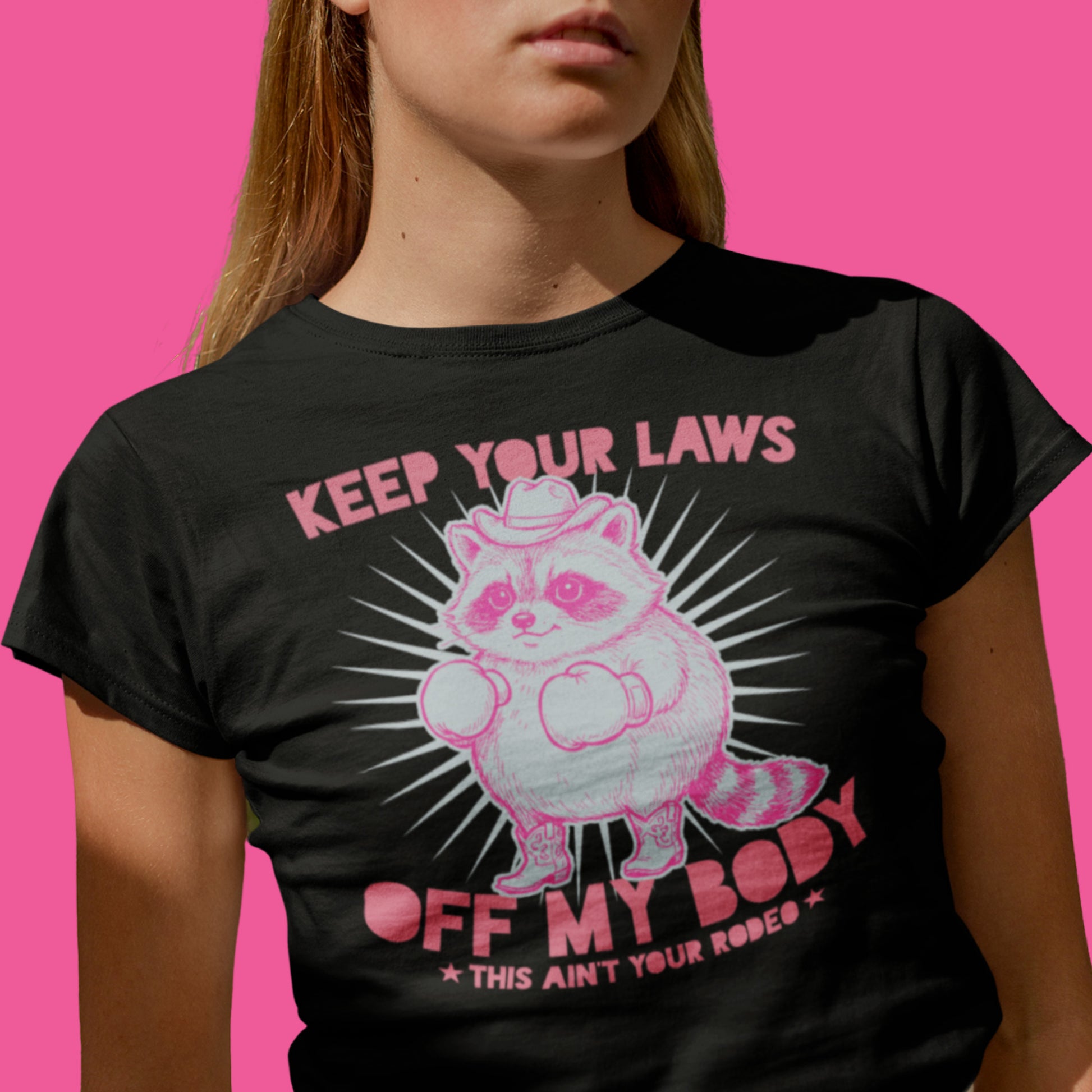 woman wearing black feminist graphic t-shirt featuring pink cowboy raccoon illustration with “Keep Your Laws Off My Body” slogan against bright pink background.