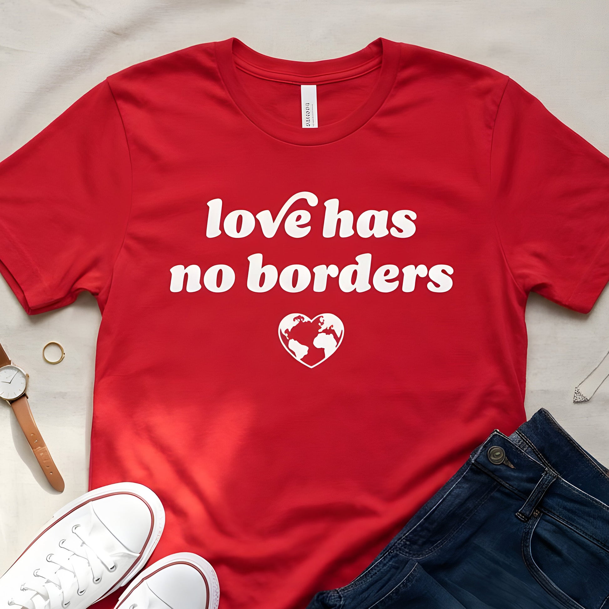 Red unisex t-shirt flat lay with jeans and sneakers, featuring bold white typography and globe heart graphic reading “love has no borders,” a justice-focused design.