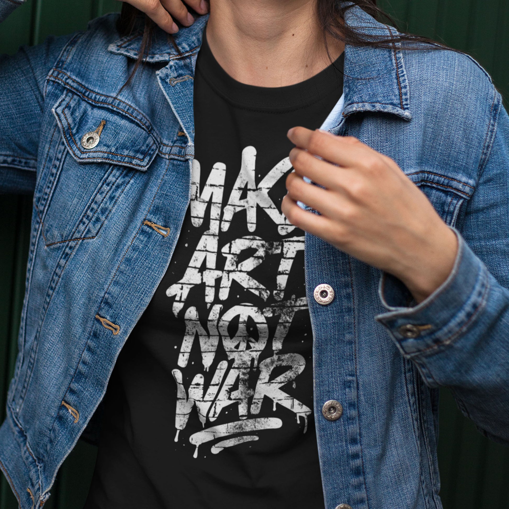 Person wearing a black Make Art Not War shirt under a denim jacket, hand adjusting the collar. A modern, effortless look from Blue Wave USA that blends art and protest culture.