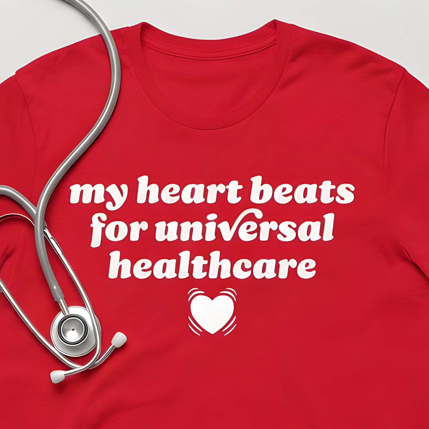 Red unisex t-shirt flat lay featuring stethoscope and bold typography reading “my heart beats for universal healthcare,” a clean healthcare solidarity graphic and justice-focused design.