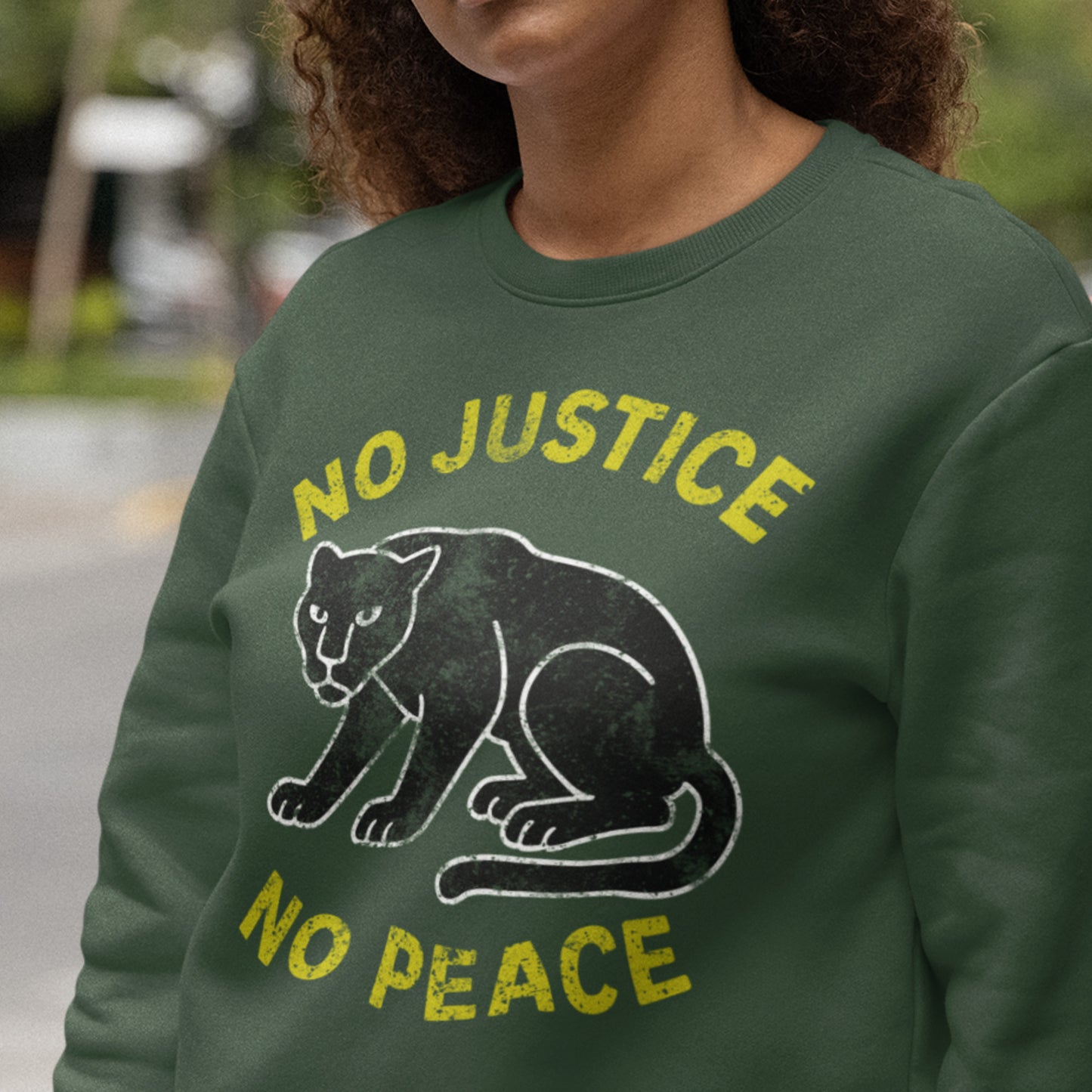 Model wearing a green unisex sweatshirt featuring “NO JUSTICE NO PEACE” and chalk-line panther artwork. Progressive clothing expressing solidarity and justice-forward advocacy.