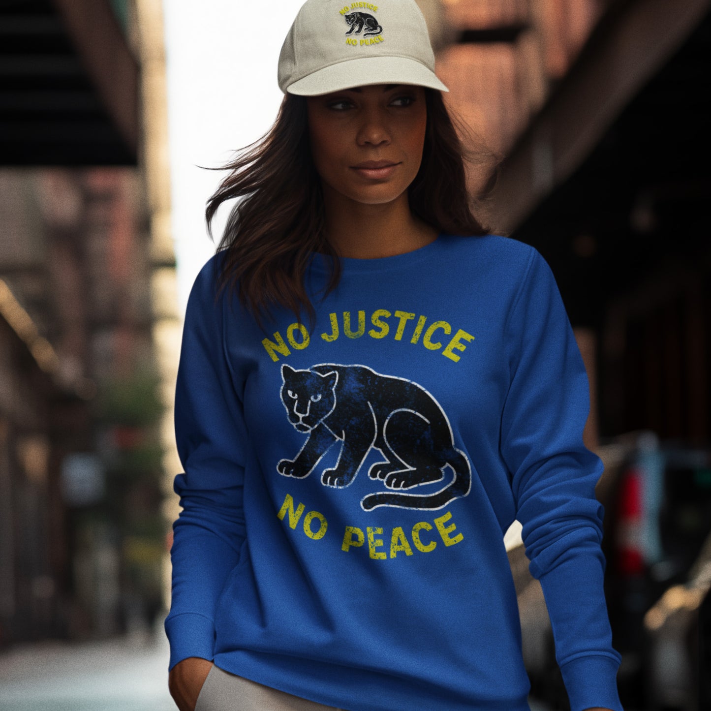 Model wearing a blue unisex sweatshirt printed with “NO JUSTICE NO PEACE” and a textured panther graphic. Activist sweatshirt styled for streetwear with strong justice-centered messaging.