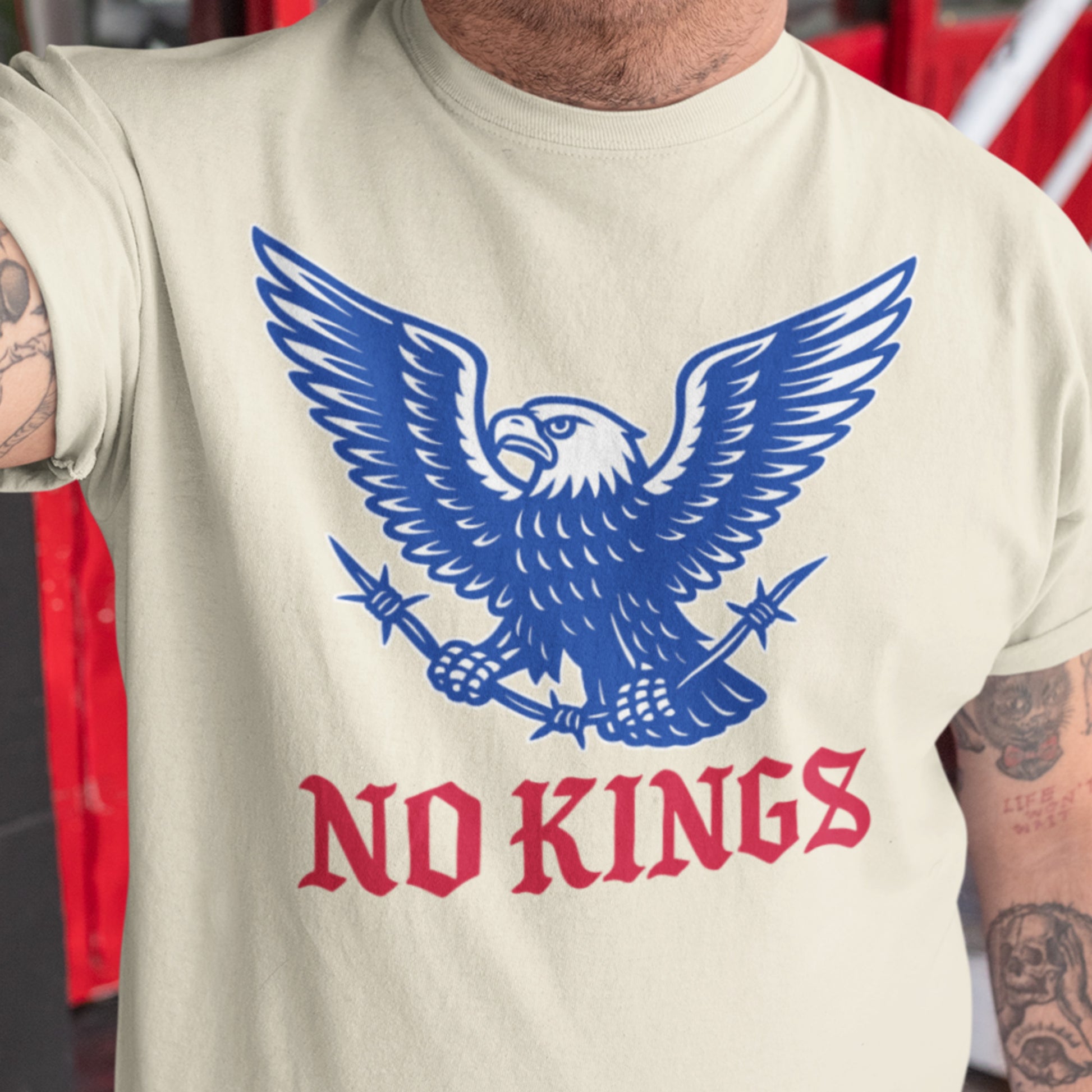 Person wearing the cream “No Kings” tee with tattooed arms visible, showing the large blue eagle graphic and red gothic lettering.