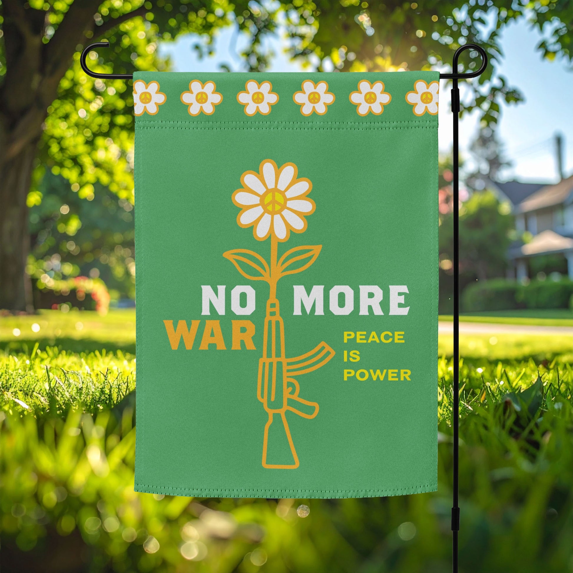 Garden flag featuring yellow daisy growing from rifle graphic with bold “NO MORE WAR” and smaller “PEACE IS POWER” message displayed on lawn stake outdoors.