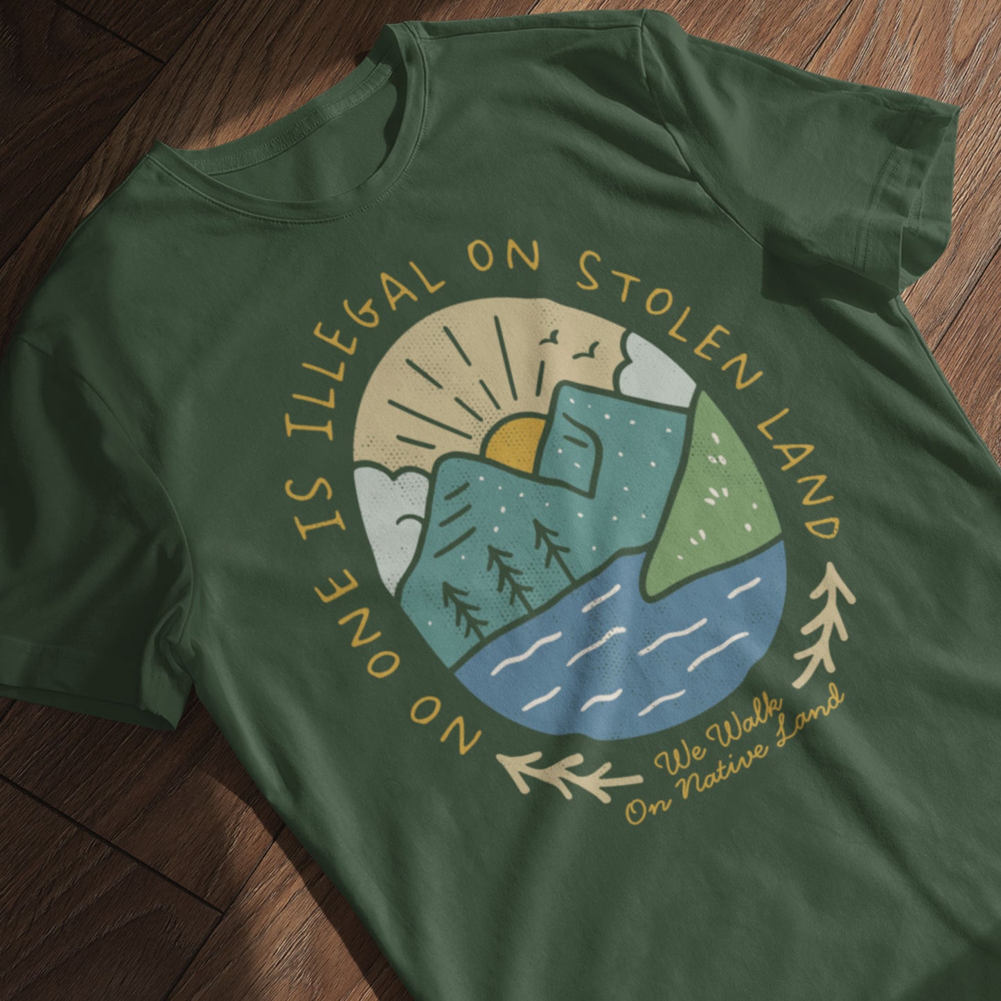 Forest Green “No One Is Illegal on Stolen Land” t-shirt folded neatly, showing the sun, river, and mountain graphic with warm earth-tone colors by Blue Wave USA.