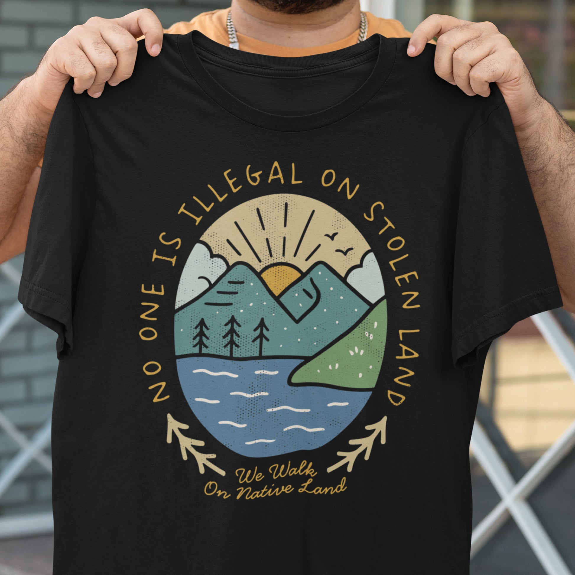Person holding up a black t-shirt featuring a circular mountain and river design with the text “No One Is Illegal on Stolen Land” and “We Walk on Native Land.” A Blue Wave USA design honoring Indigenous sovereignty and solidarity.