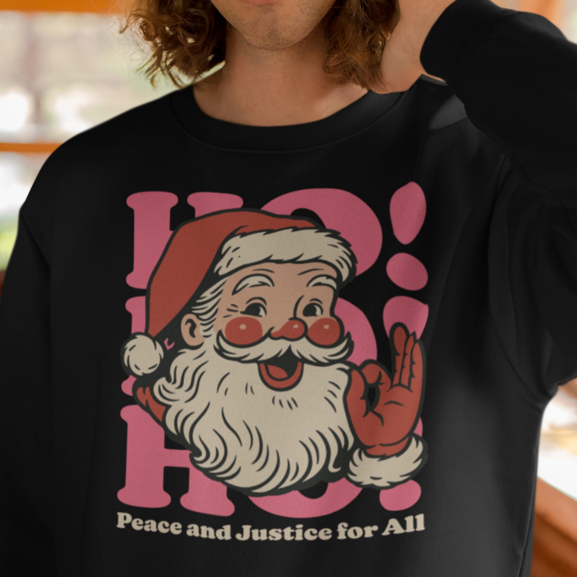 Model wearing a black sweatshirt with a vintage Santa illustration, pink “HO!” layout, and the printed message “Peace and Justice for All.”