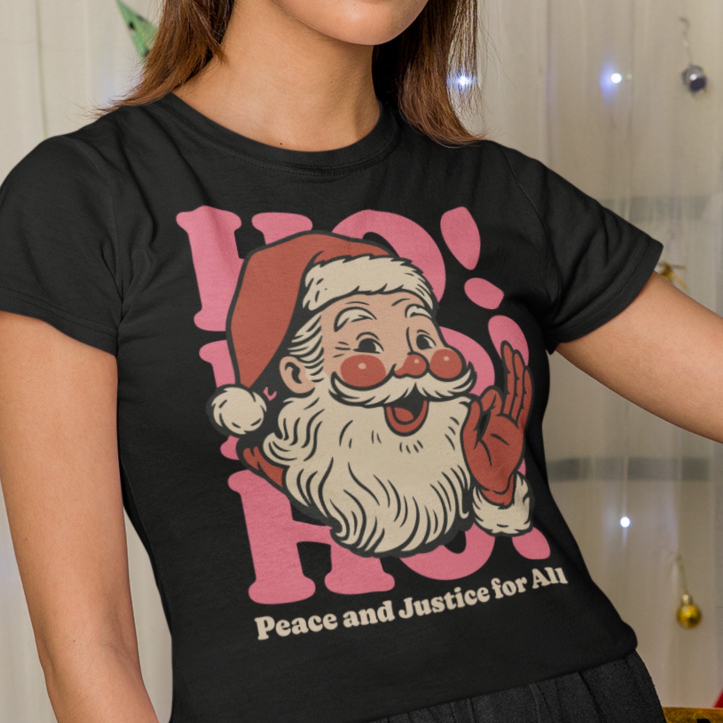 Woman wearing a black Christmas t-shirt with a vintage Santa and “Peace and Justice for All” design, posing in front of holiday lights.