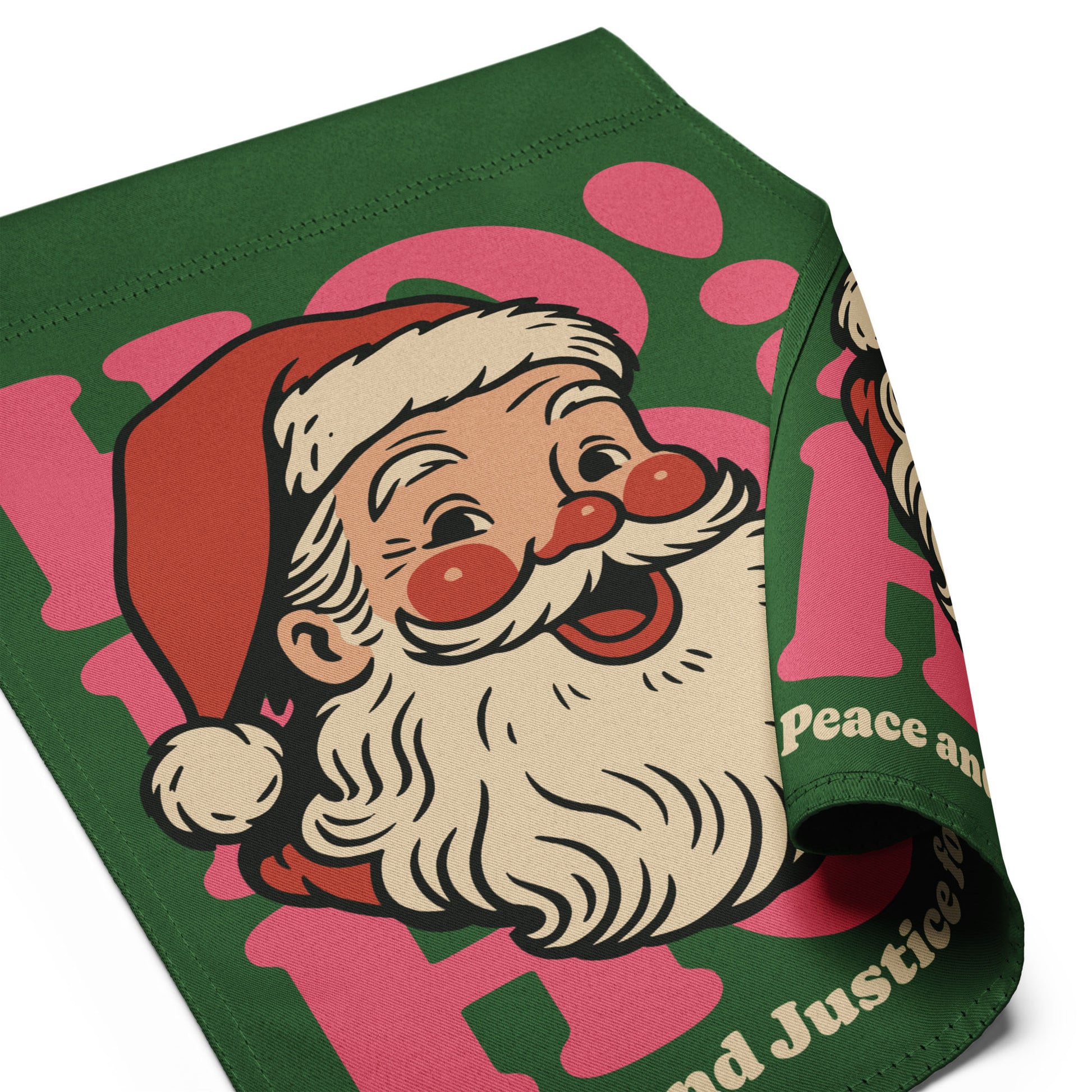 Close-up of a rolled green garden flag featuring a vintage Santa illustration with pink “HO!” accents and the printed phrase “Peace and Justice for All.”