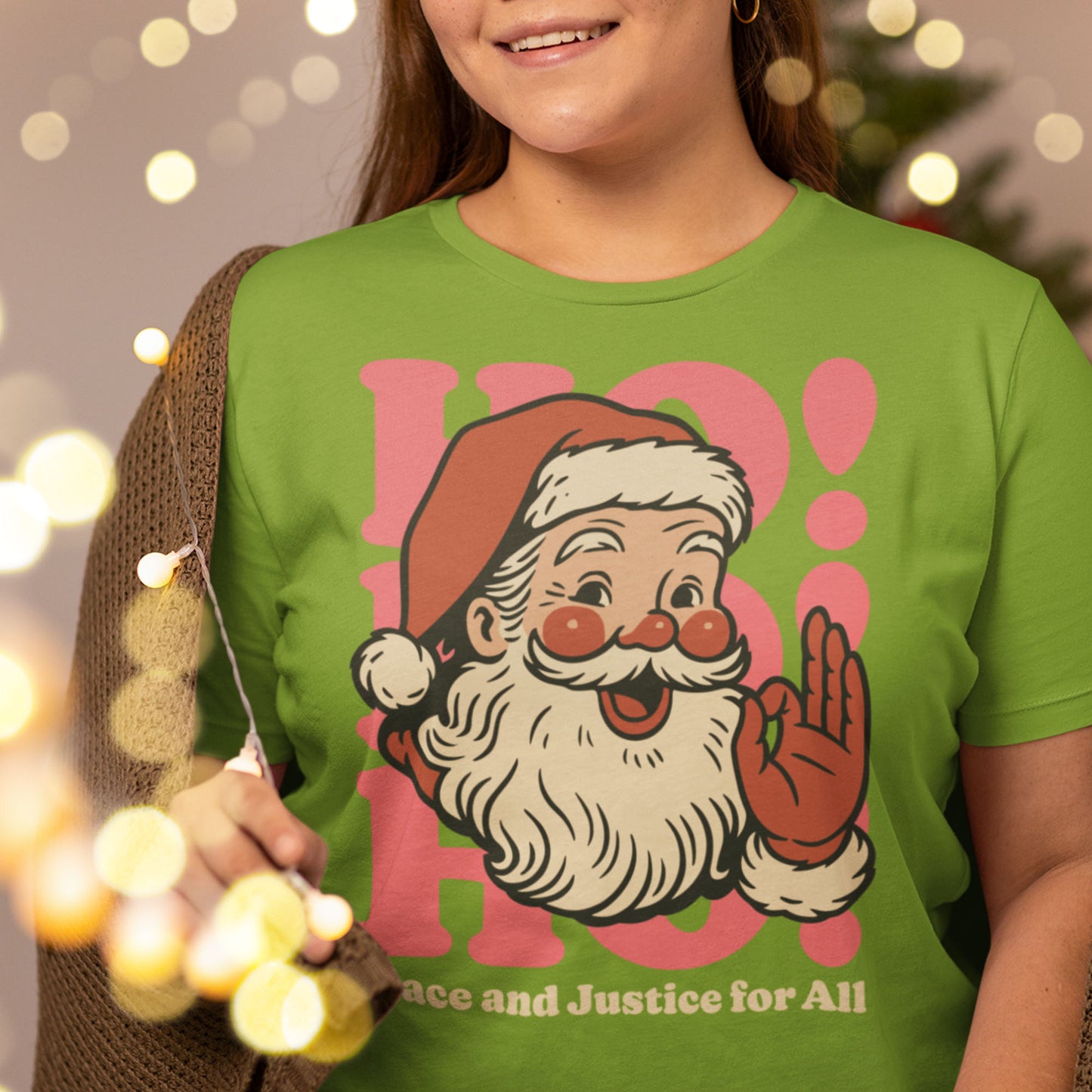 Person wearing an leaf green Santa “Peace and Justice for All” t-shirt, photographed with warm holiday bokeh lights.