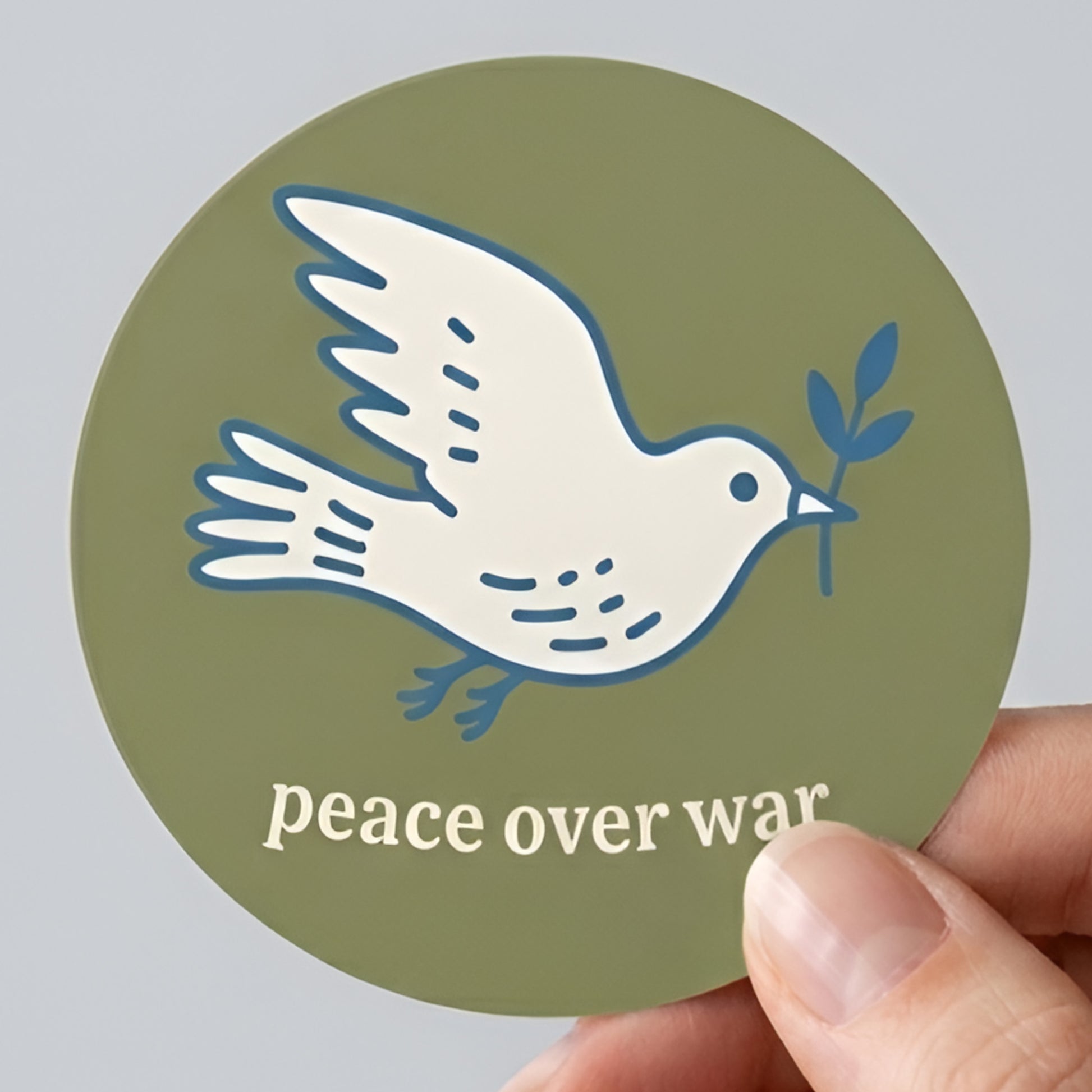 Round sticker featuring a white dove holding an olive branch on olive green background with “peace over war” text, minimalist anti-war activist design.