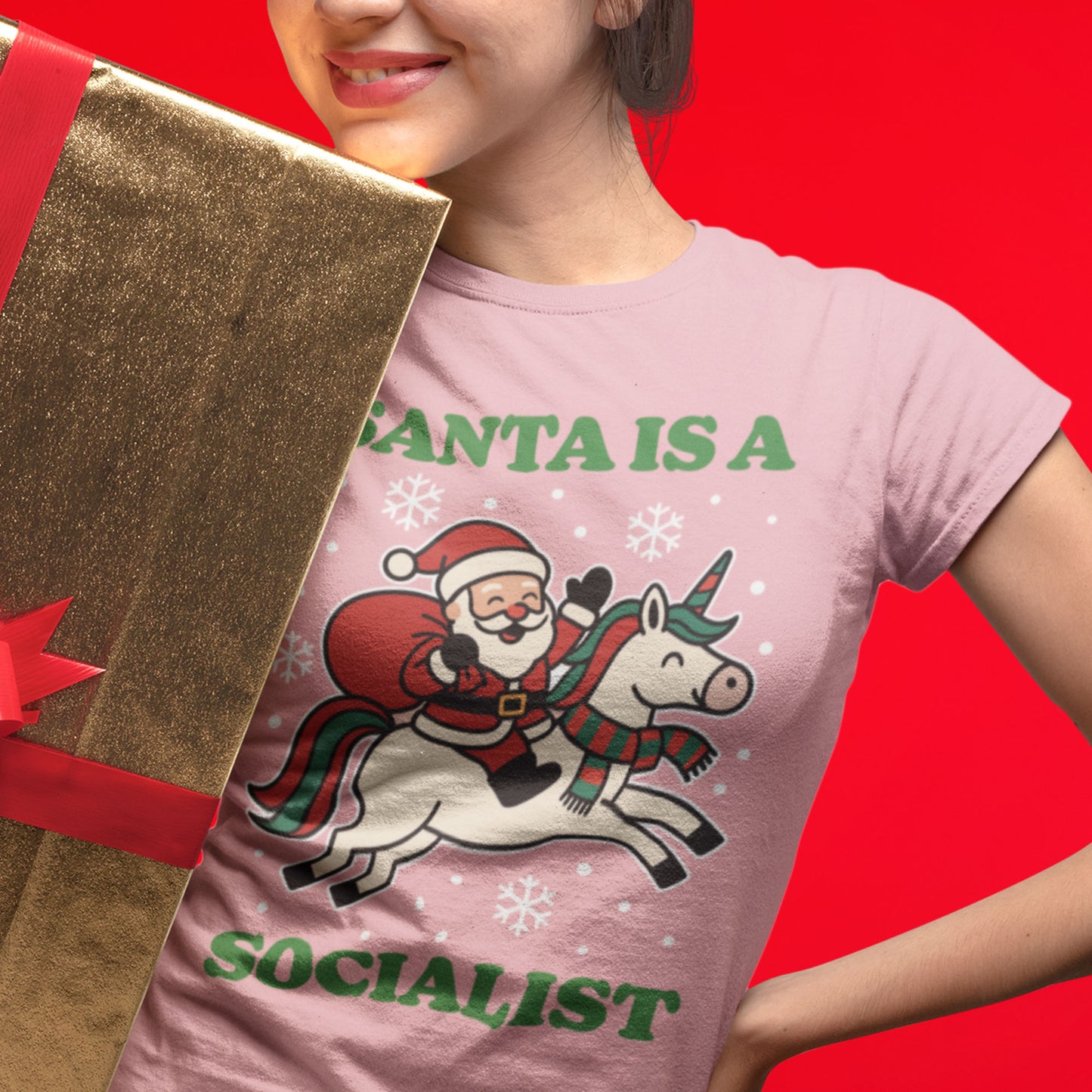 Model holding a gold-wrapped gift while wearing a pink women’s relaxed t-shirt featuring a retro Santa riding a unicorn with stars and the printed phrase “SANTA IS A SOCIALIST.”