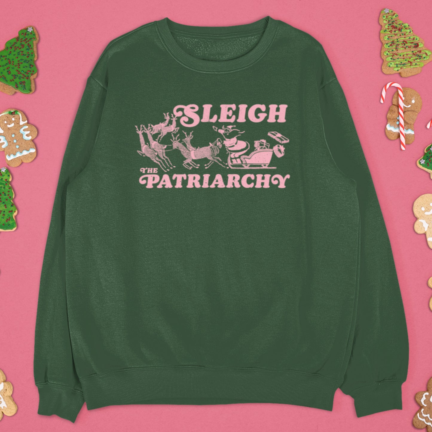 Green sweatshirt with the pink “Sleigh the Patriarchy” design displayed on a pink festive backdrop with ornaments and ribbon.