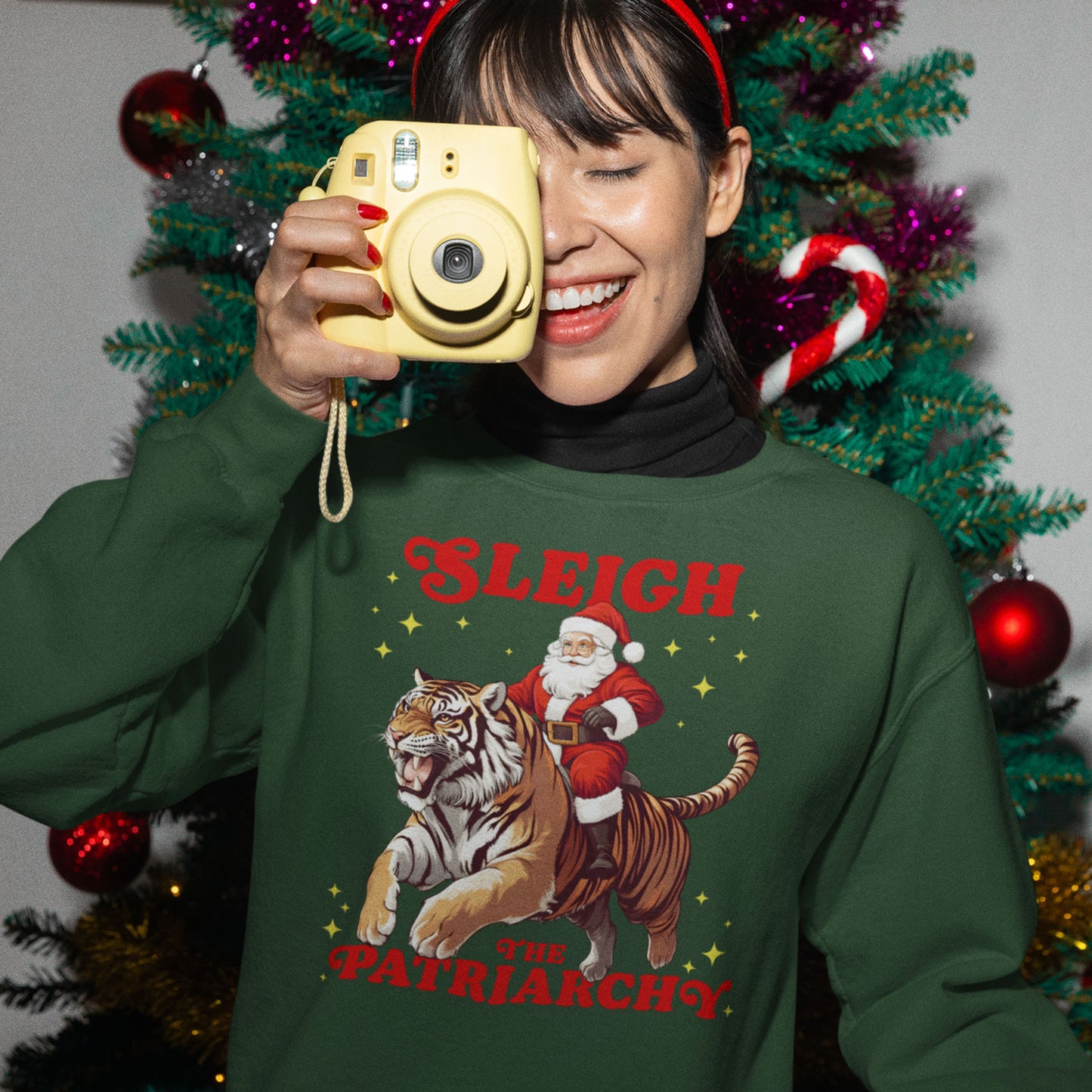 Person smiling and holding a camera while wearing a green “Sleigh the Patriarchy” sweatshirt with Santa riding a tiger. Festive lights and ornaments in the background. A spirited holiday design from Blue Wave USA.