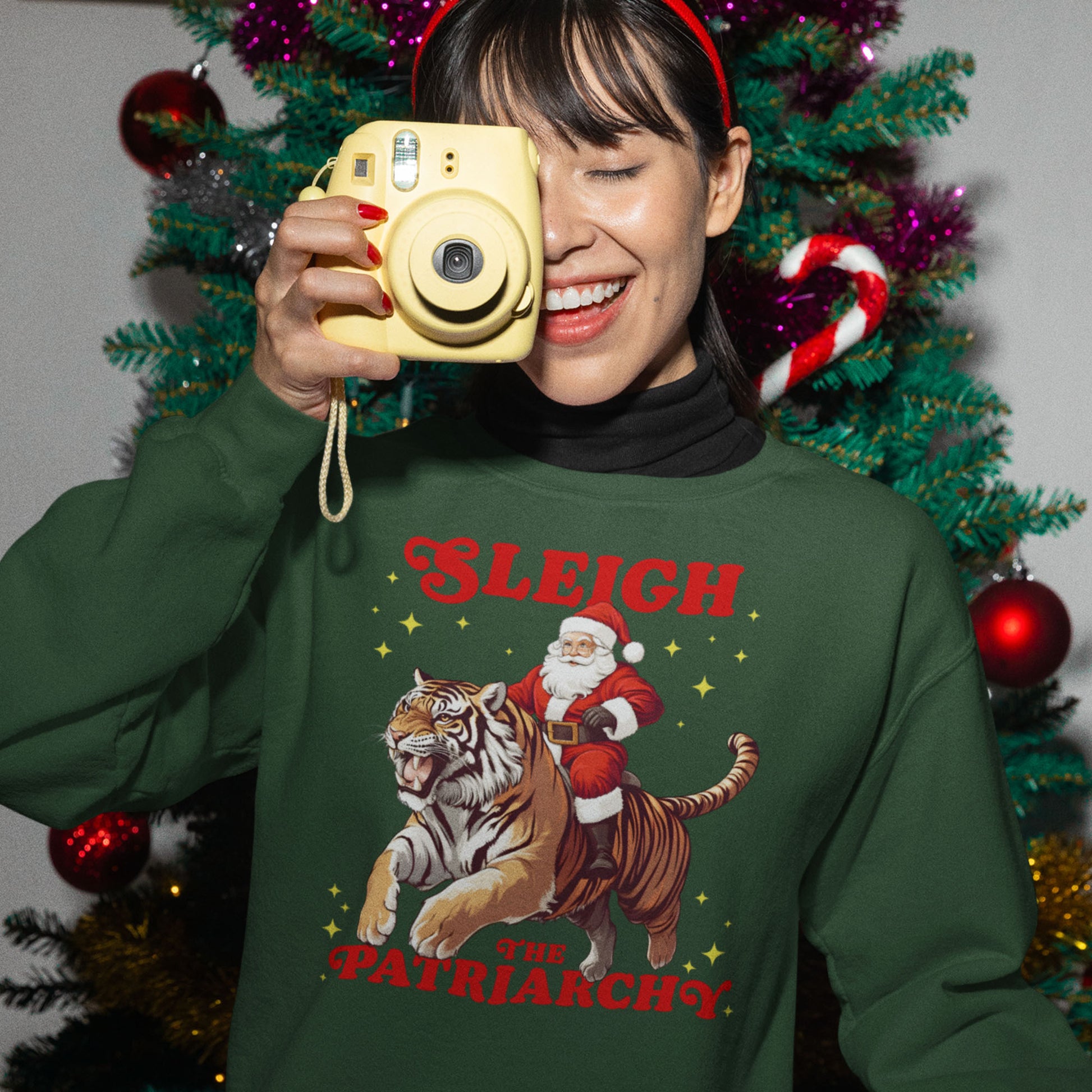 Person smiling and holding a camera while wearing a green “Sleigh the Patriarchy” sweatshirt with Santa riding a tiger. Festive lights and ornaments in the background. A spirited holiday design from Blue Wave USA.