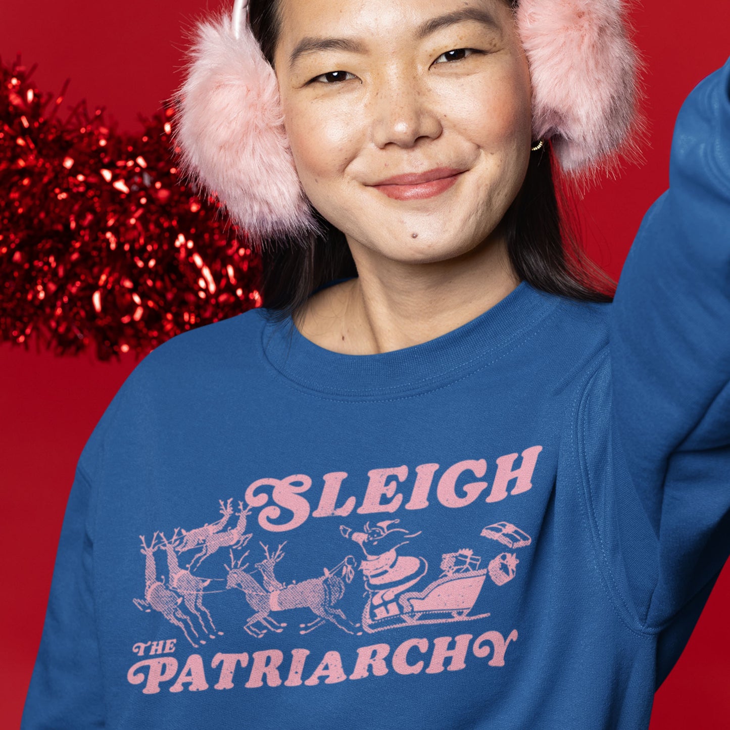 Person smiling in a blue sweatshirt with the pink “Sleigh the Patriarchy” graphic, standing in front of festive red holiday decor. A playful holiday statement piece from Blue Wave USA.