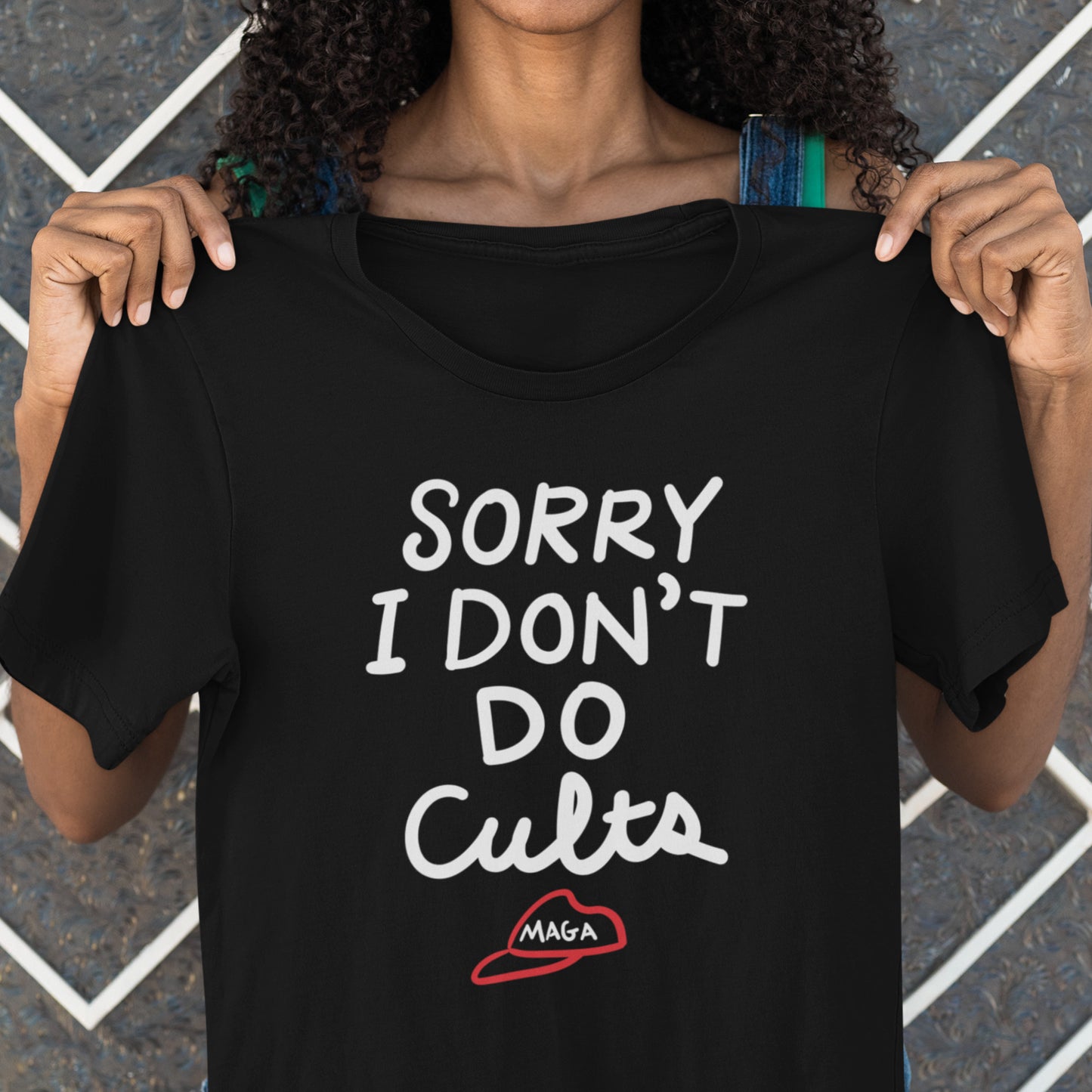 Person holding black unisex t-shirt showing bold white lettering reading “SORRY I DON’T DO CULTS.” with crossed-out MAGA hat symbol.