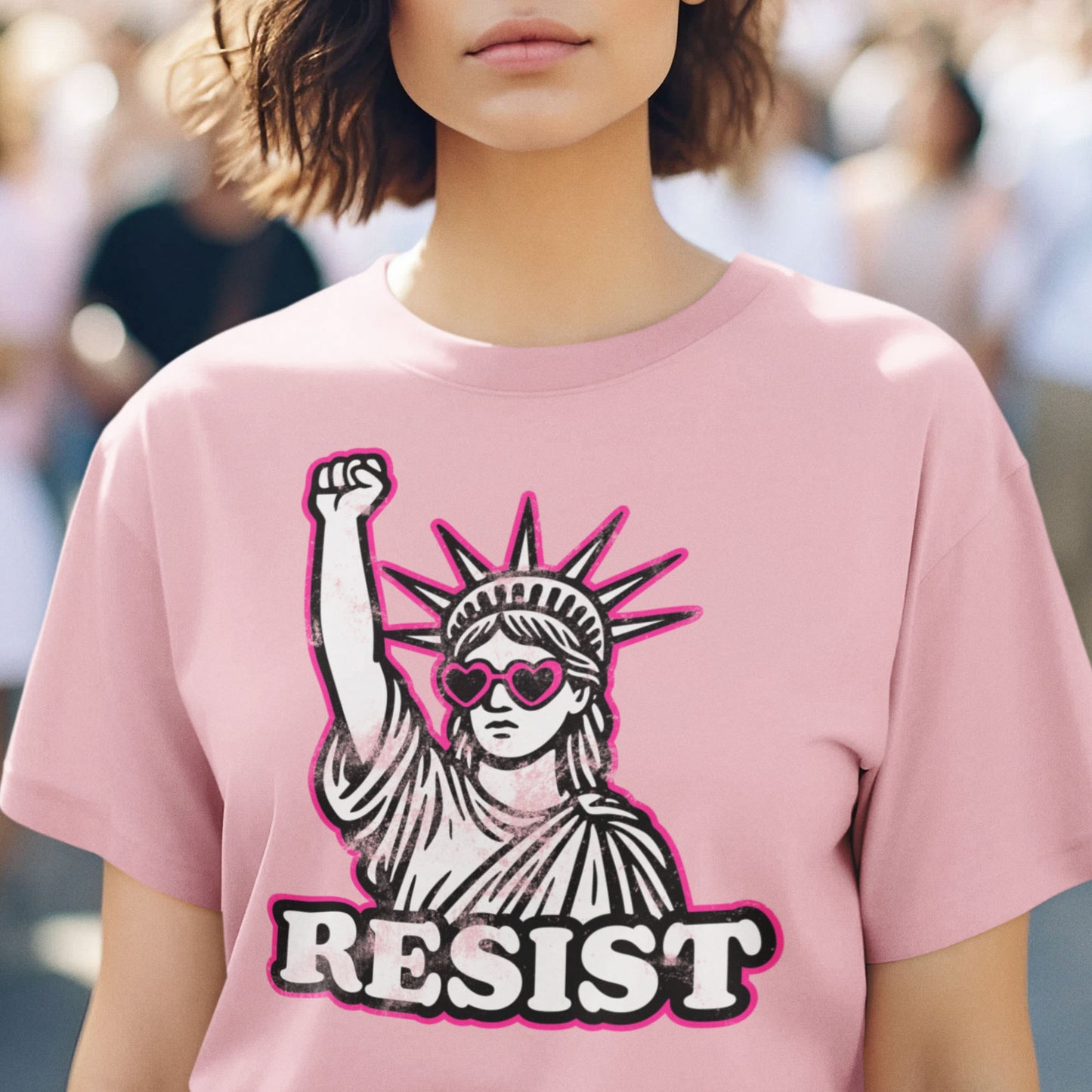 Person wearing a soft pink “RESIST” t-shirt with the raised-fist Statue of Liberty graphic, representing strength, equality, and collective action.