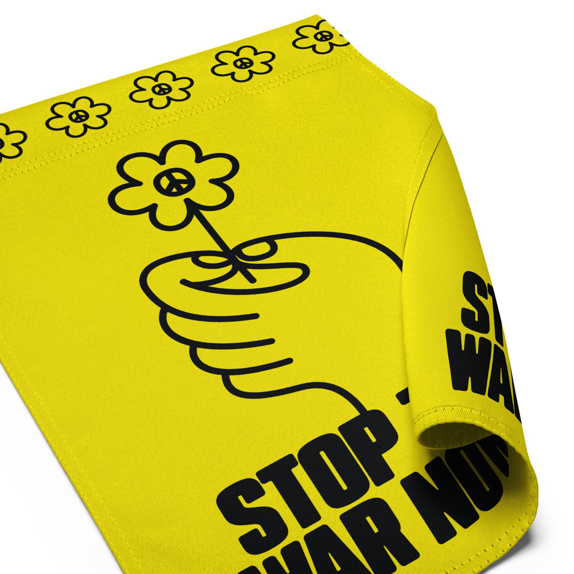 Close-up of yellow garden flag fabric showing decorative flower border along top and partial “STOP THE WAR NOW!” text.