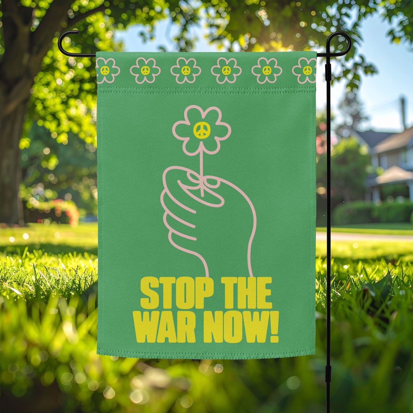 Green garden flag featuring hand holding flower illustration with bold “STOP THE WAR NOW!” message displayed on yard stake in front lawn setting.