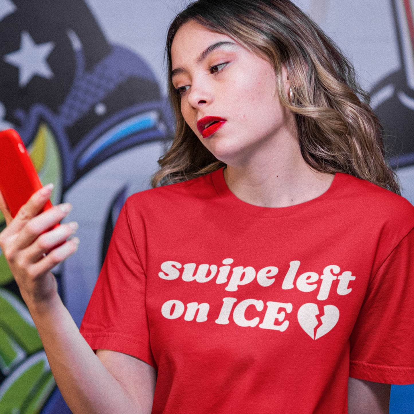 Unisex red t-shirt worn by a person holding a phone, featuring bold white typography and heart graphic with the phrase “swipe left on ICE,” styled for progressive Valentine messaging.