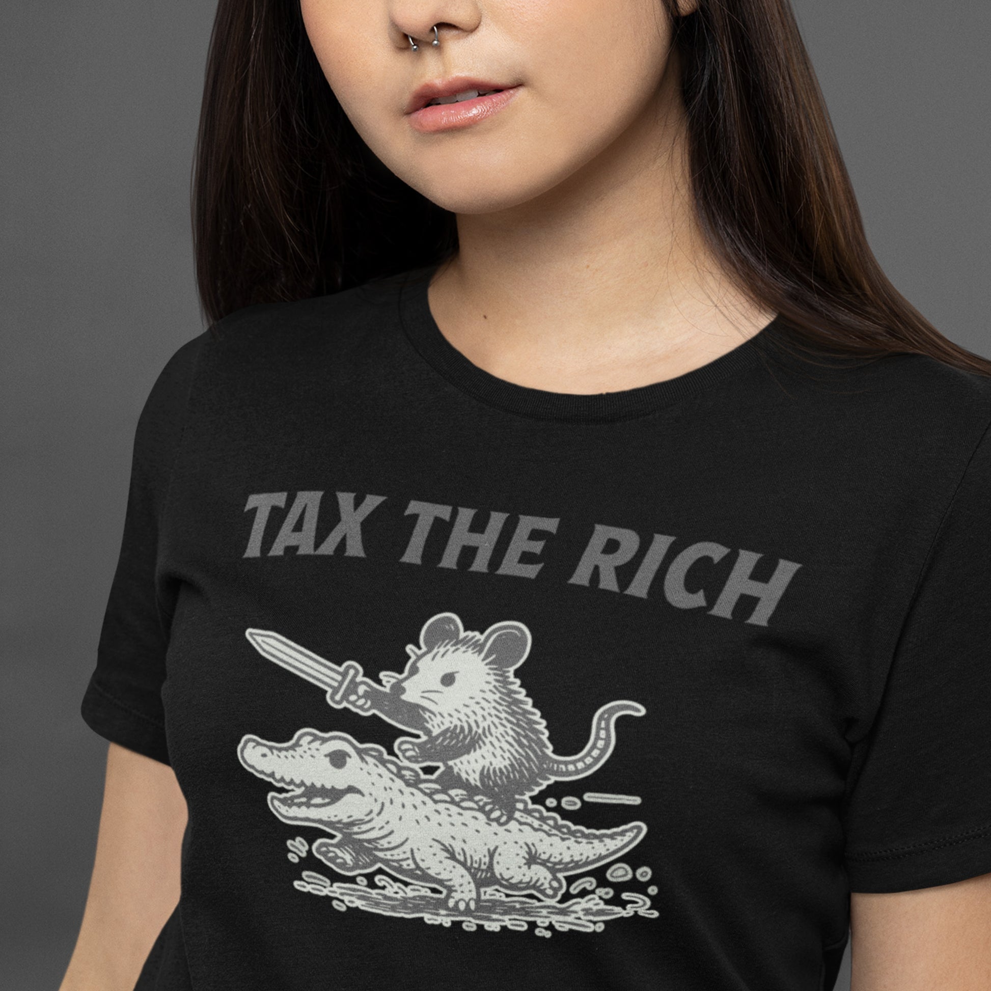 Person wearing a black unisex t-shirt featuring a vintage-style opossum riding an alligator while holding a sword beneath the phrase “Tax the Rich,” a humorous anti-billionaire graphic inspired by protest art, economic justice, and political satire.
