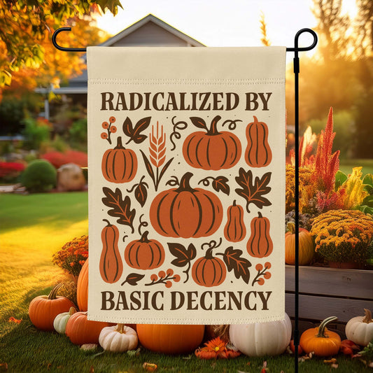 “Radicalized By Basic Decency” Thanksgiving garden flag with folk-art pumpkins and autumn leaves, displayed outdoors among fall décor.