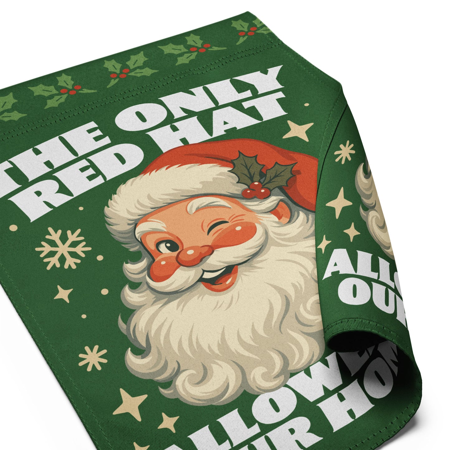 Close-up of a partially rolled green garden flag featuring a retro Santa illustration, holiday accents, and the printed message “THE ONLY RED HAT ALLOWED IN OUR HOME.”