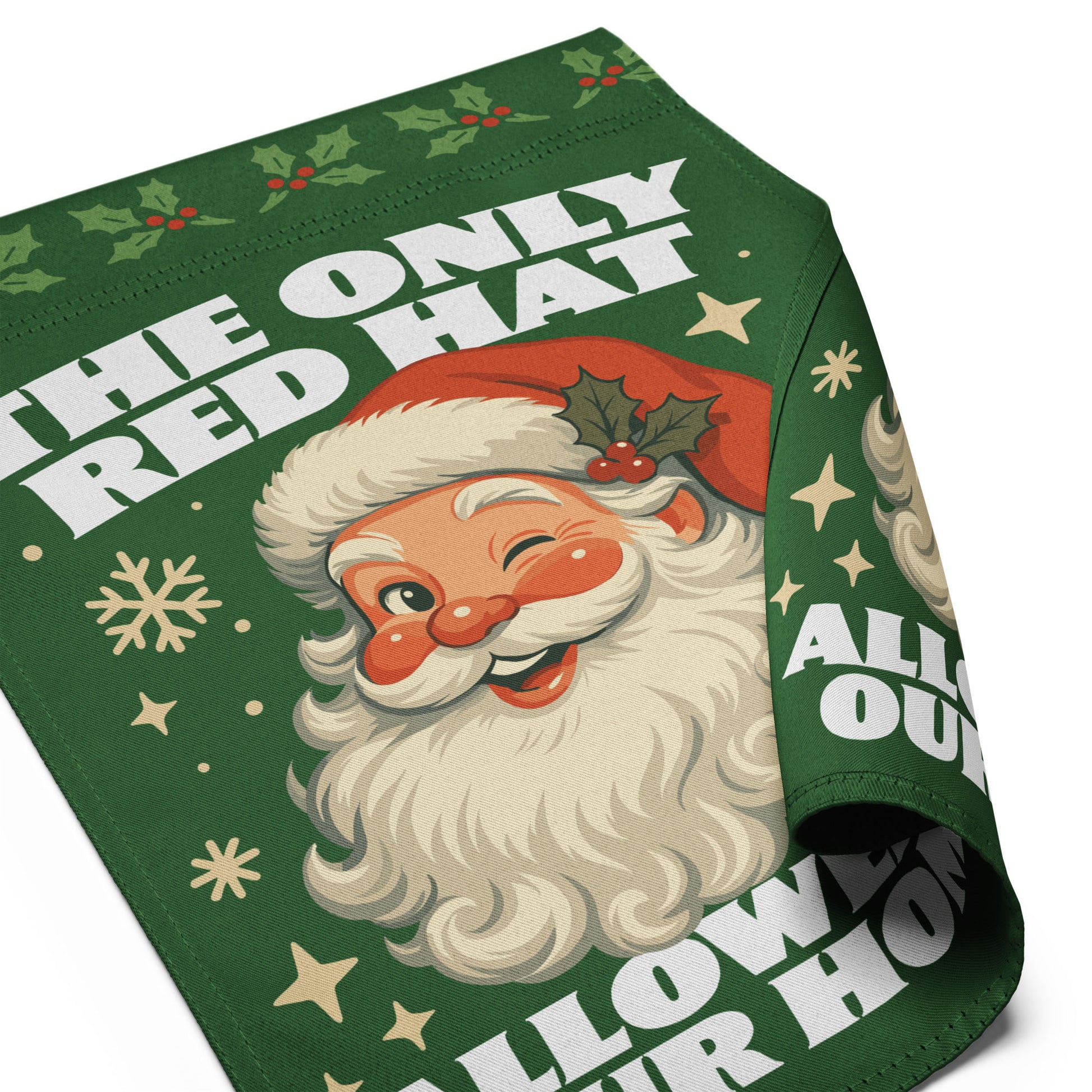 Close-up of a partially rolled green garden flag featuring a retro Santa illustration, holiday accents, and the printed message “THE ONLY RED HAT ALLOWED IN OUR HOME.”
