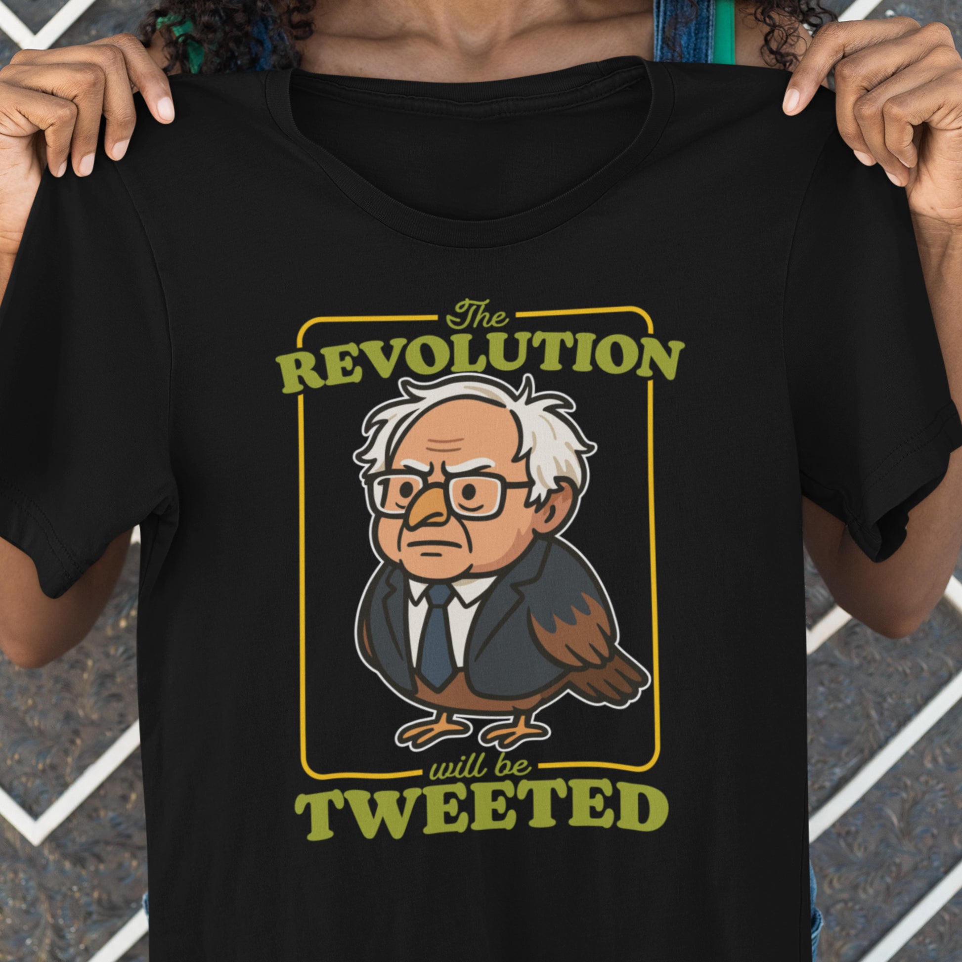 Person holding black t-shirt featuring cartoon Birdie Sanders character with glasses and “The Revolution Will Be Tweeted” text in bold retro political graphic design.