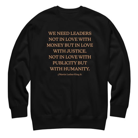 Black unisex premium sweatshirt featuring the MLK quote: “WE NEED LEADERS NOT IN LOVE WITH MONEY BUT IN LOVE WITH JUSTICE… PUBLICITY BUT WITH HUMANITY.” Clean, bold civil-rights message.