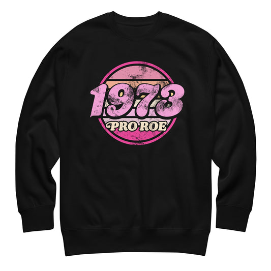 Black unisex premium sweatshirt featuring a distressed retro circular badge in pink and cream reading “1973 PRO ROE,” vintage reproductive freedom design centered on a solid black fleece.