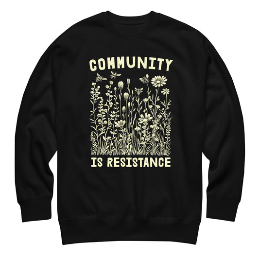 Black unisex premium sweatshirt featuring wildflower illustration with phrase “COMMUNITY IS RESISTANCE,” vintage botanical graphic, centered progressive activist apparel.