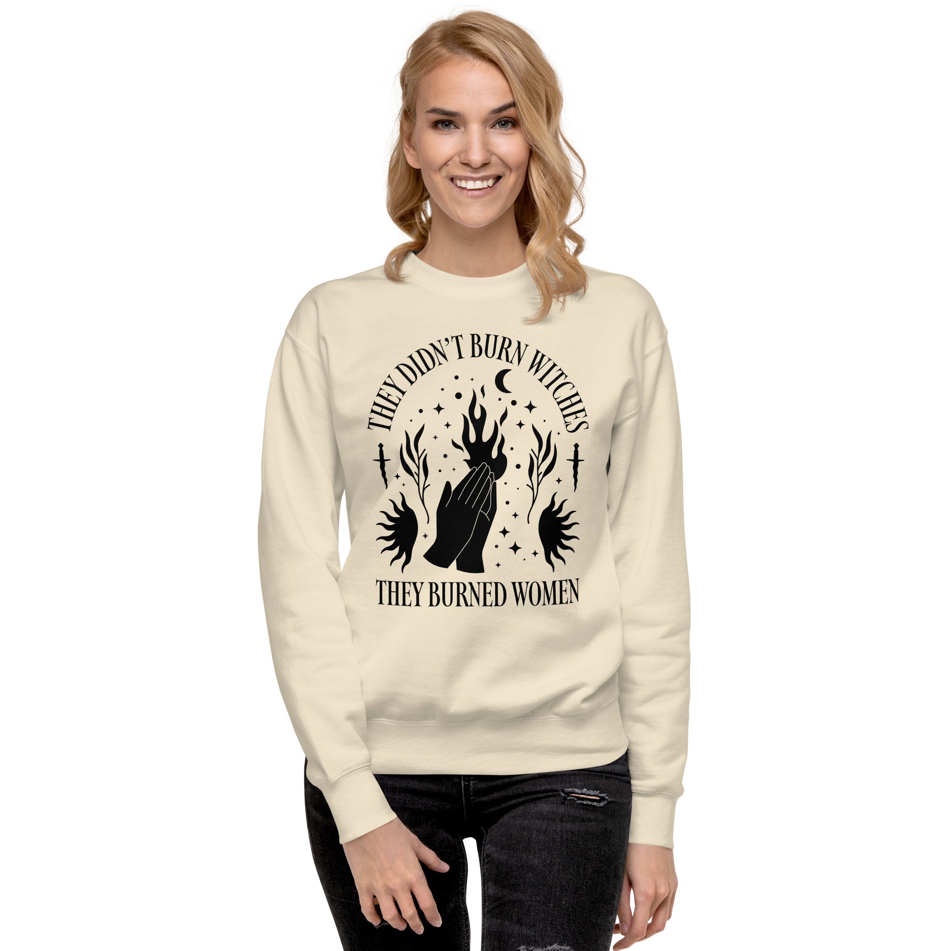 Model wearing cream unisex premium sweatshirt with mystical illustration of hands rising through flames, celestial accents, centered layout, printed message “THEY DIDN’T BURN WITCHES THEY BURNED WOMEN.”