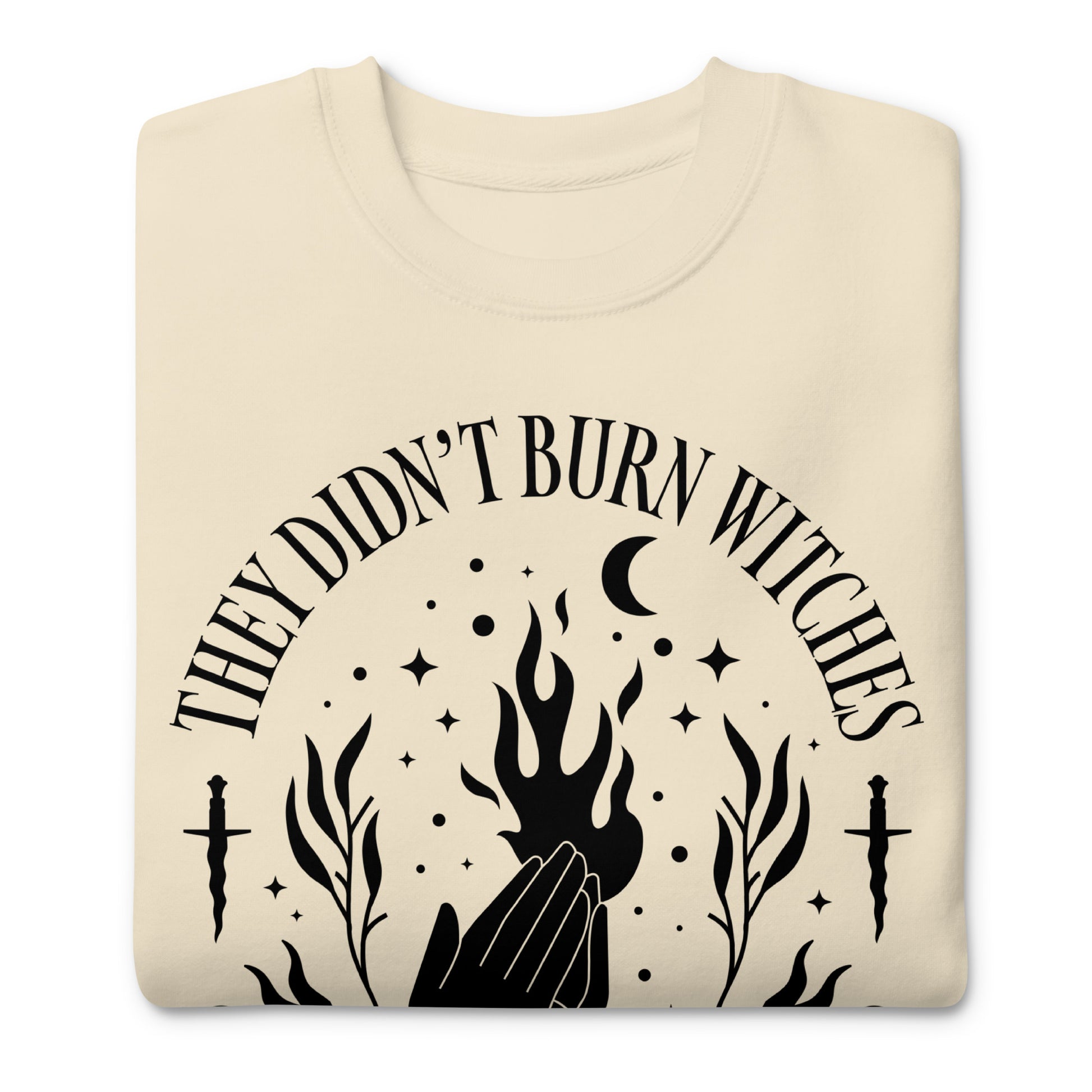 Folded cream unisex premium sweatshirt featuring bold witch-themed illustration with flames and stars, symmetrical layout, printed message “THEY DIDN’T BURN WITCHES THEY BURNED WOMEN,” feminist apparel from Blue Wave USA.