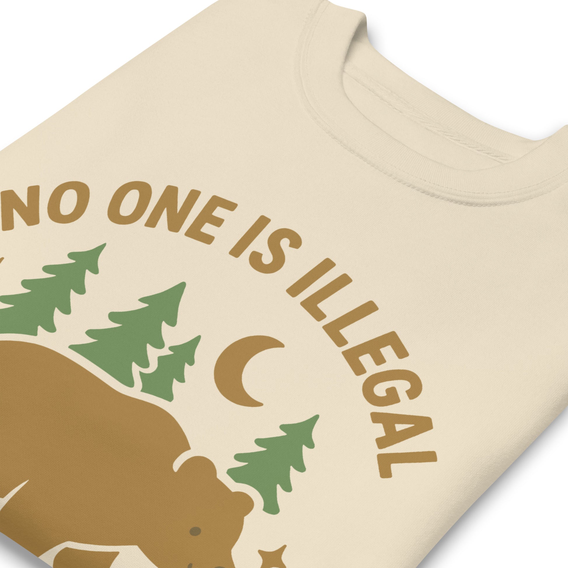 Close-up of a cream unisex premium sweatshirt highlighting the bear, trees, and moon graphic and the message “NO ONE IS ILLEGAL ON STOLEN LAND,” shown in warm earthy print tones.
