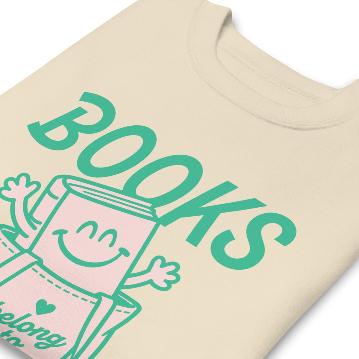 Close-up of the “Books Belong to Everyone” design on a cream sweatshirt, highlighting the playful illustrated book character and teal typography.