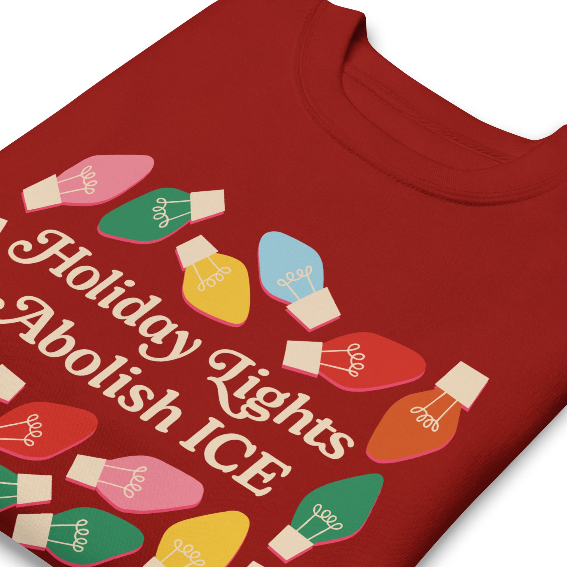 A close-up of a red unisex sweatshirt showing bright multicolor holiday bulbs framing the message “Holiday Lights Abolish ICE,” emphasizing the vibrant seasonal activism artwork.
