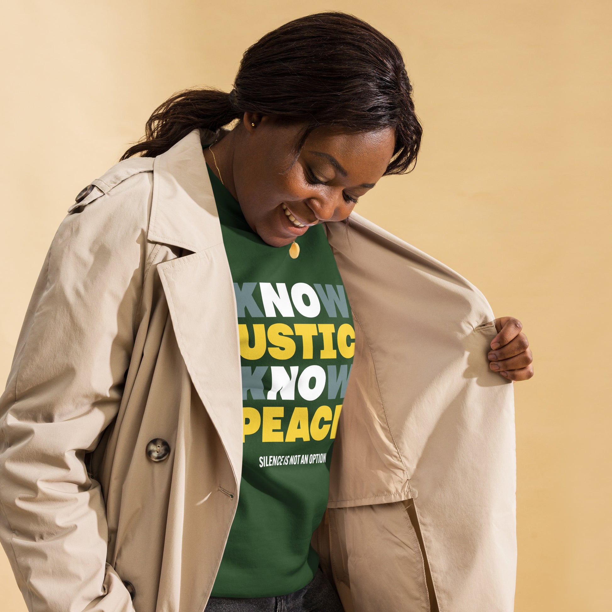 Person wearing a forest green Know Justice Know Peace sweatshirt under a tan trench coat, slightly turned and smiling — showing activism and everyday style coming together. Designed by Blue Wave USA for those who lead with conscience.