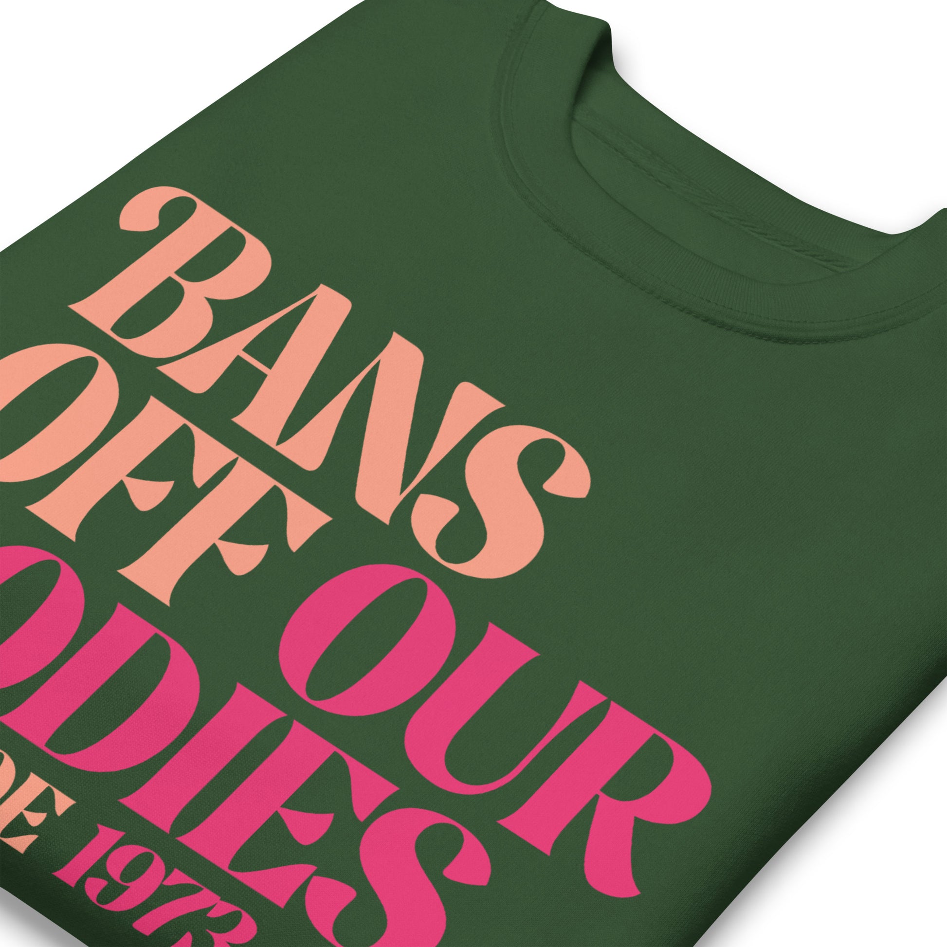 Close-up of forest green unisex premium sweatshirt showing retro serif lettering in pink gradient, stacked typographic layout, printed message “BANS OFF OUR BODIES PRO ROE 1973.”