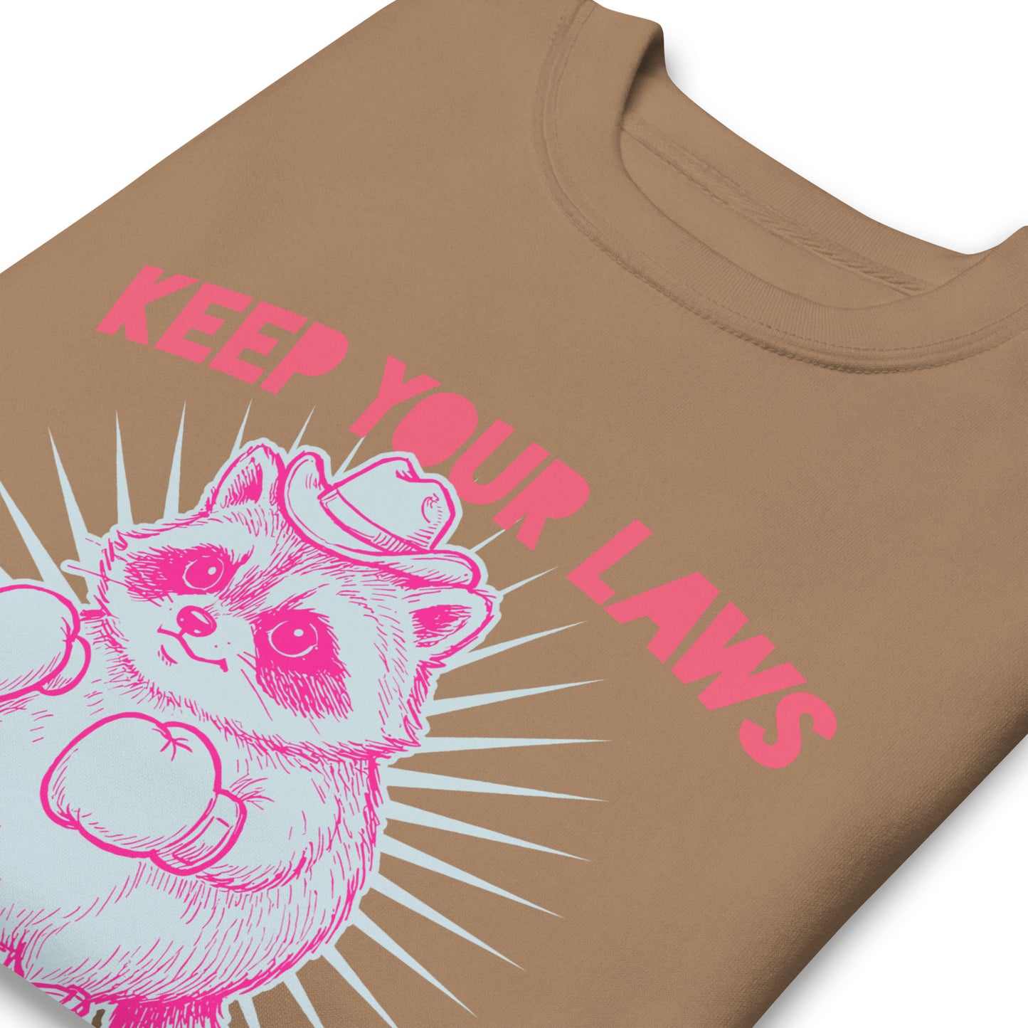 Close-up latte brown sweatshirt print showing pink raccoon cowboy illustration with “Keep Your Laws” wording in western feminist activist design.
