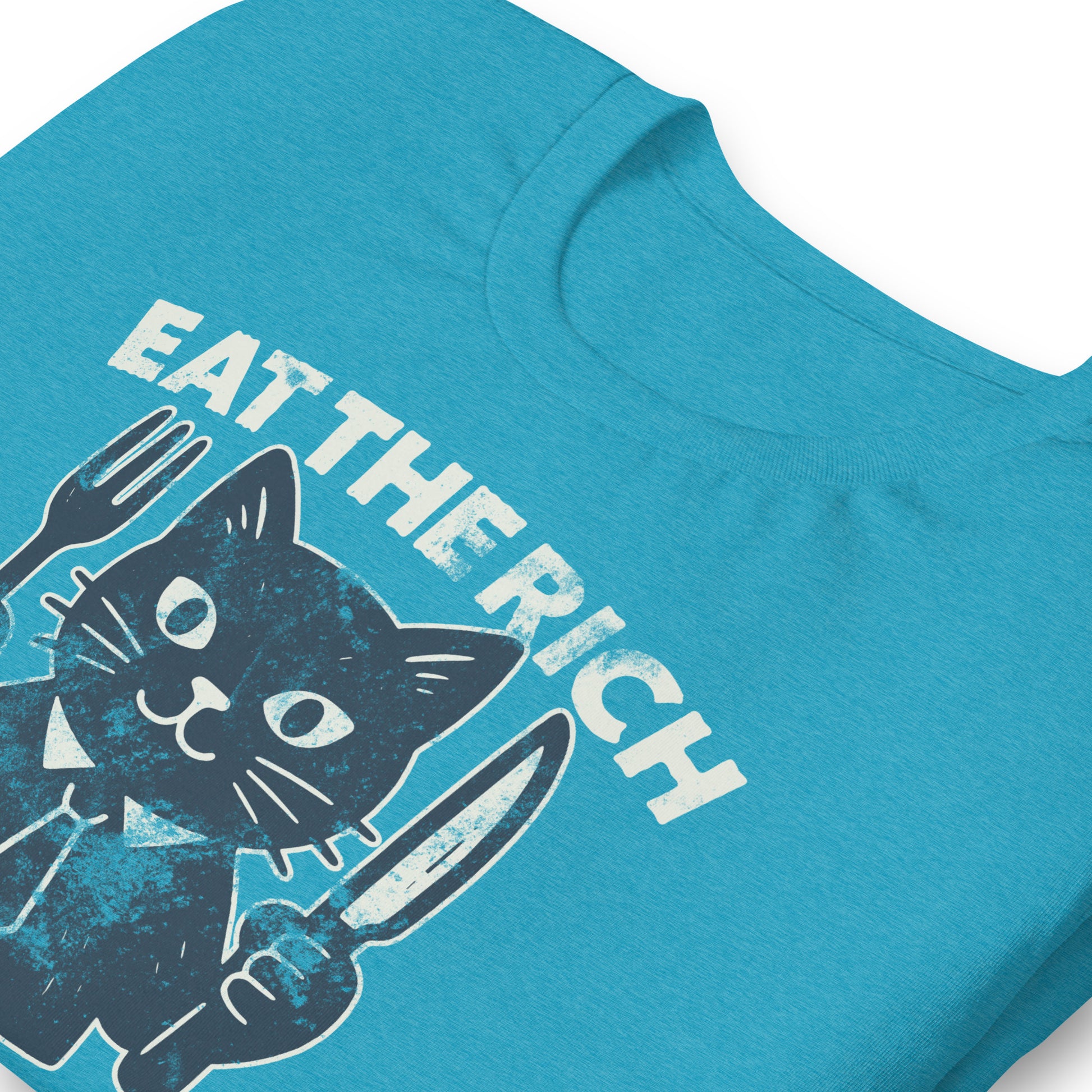 Folded heather aqua t-shirt displaying a playful cat illustration holding cutlery beneath the phrase “Eat the Rich,” combining political humor and protest-inspired design.