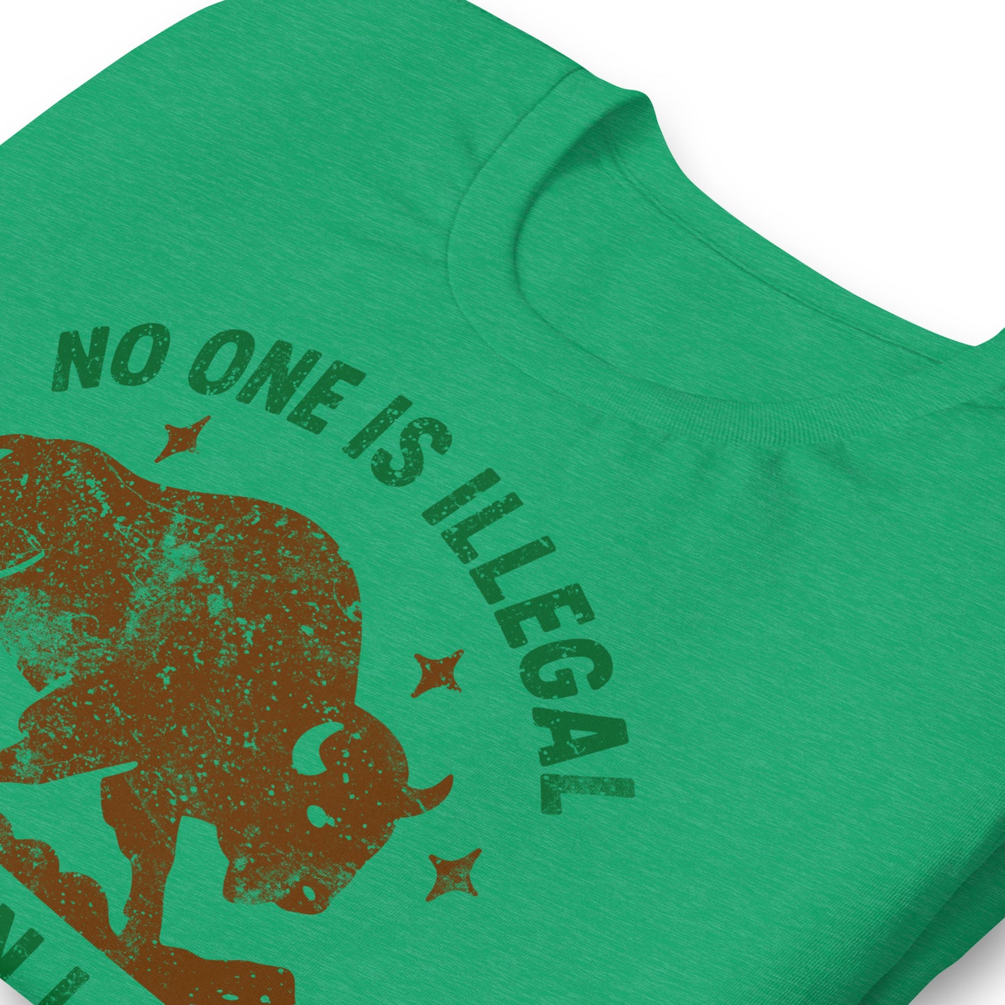 Close-up view of a heather kelly green t-shirt showing part of the buffalo artwork and the message “NO ONE IS ILLEGAL ON STOLEN LAND,” highlighting texture and earthy print details.