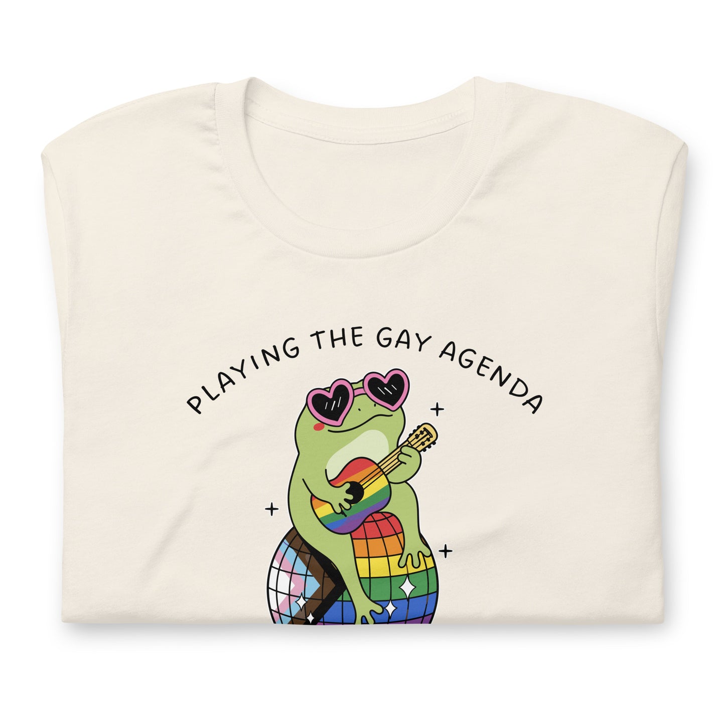Folded vintage white pride t-shirt displaying a disco frog illustration with rainbow details, heart-shaped sunglasses, and playful “Playing the Gay Agenda” LGBTQ+ design.