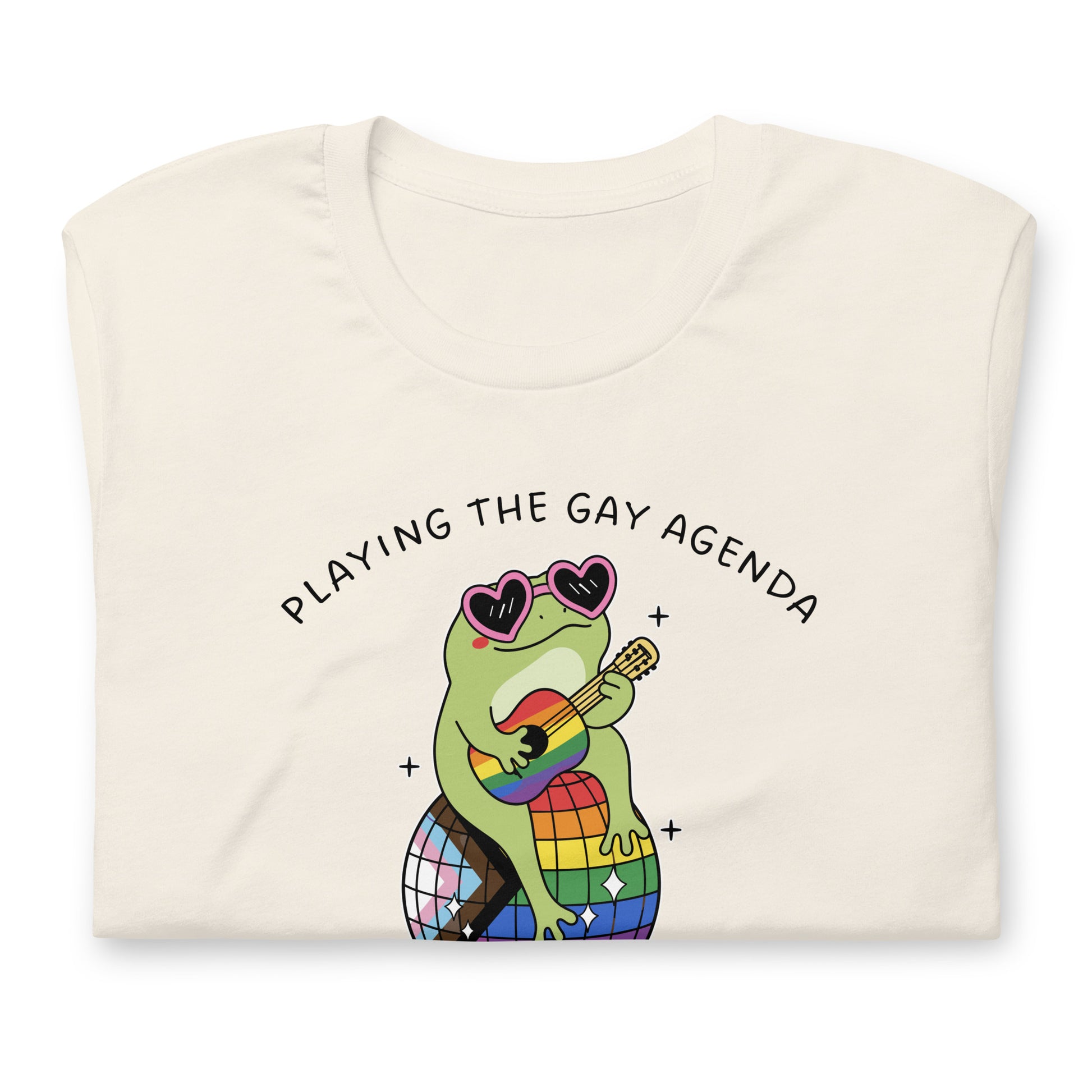 Folded vintage white pride t-shirt displaying a disco frog illustration with rainbow details, heart-shaped sunglasses, and playful “Playing the Gay Agenda” LGBTQ+ design.