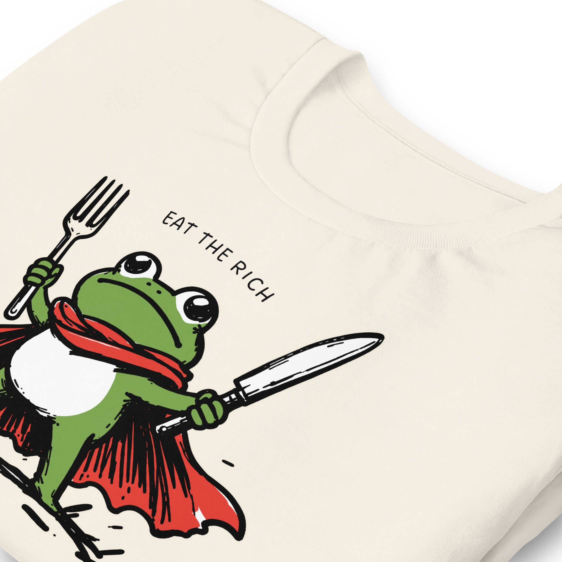 Close-up of a vintage white t-shirt featuring a cartoon hero frog with fork and knife, cape details, and the phrase “Eat the Rich.”
